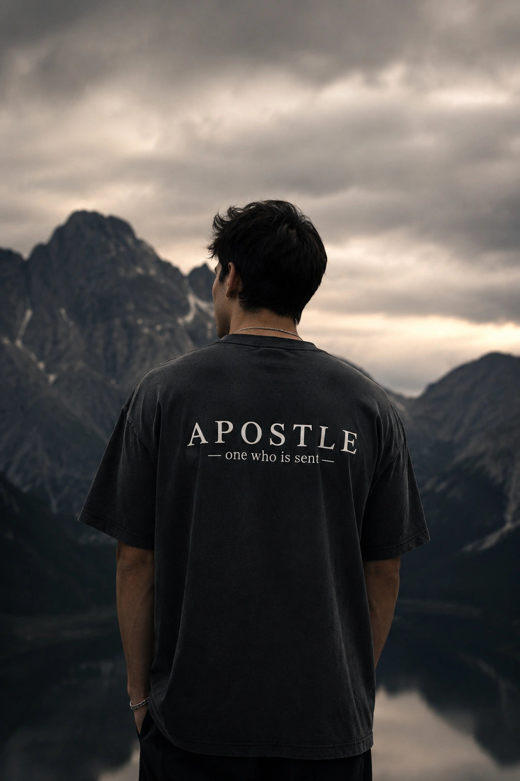 A man viewed from behind wearing a dark charcoal gray t-shirt with 'APOSTLE - one who is sent-' printed in white across the back, set against a dramatic mountain and lake landscape under a cloudy sky.